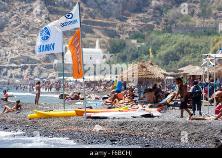 Santorini, Thira. Telephoto shot along Kamari beach with rows of sun recliners, parasols, with sunbathers and tourists, rocky cliff face as background. Stock Photo