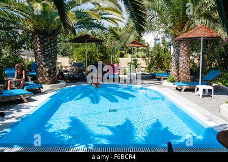 Santorini, Thira. Hotel Hermes swimming pool. Palm trees at end and woman in red bikini sitting on edge of pool, facing viewer. Palm trees above her. Stock Photo