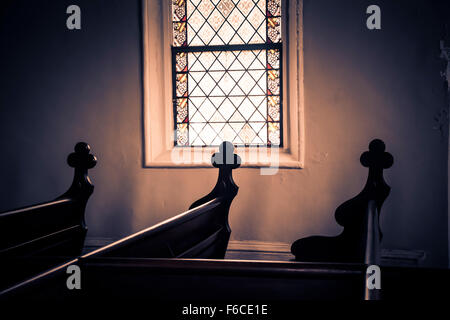 A dimly lit interior from a stained glass church window with carved crosses decoratively set on the ends of each wooden pew art Stock Photo