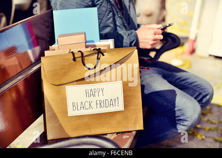 closeup of a young caucasian man sitting in a bench using his smartphone with some shopping bags, one of them with the text happ Stock Photo