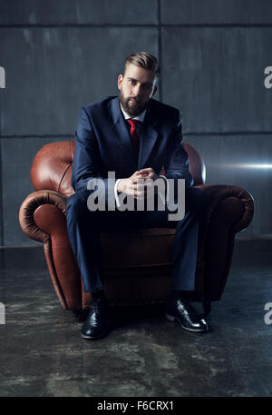 Young handsome businessman with beard in black suit sitting on chair and looking on camera. Stock Photo