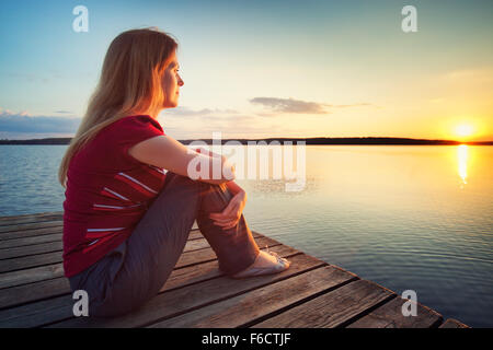Young woman in casual clothing sitting on wooden bridge and looking on sunset over lake. Stock Photo