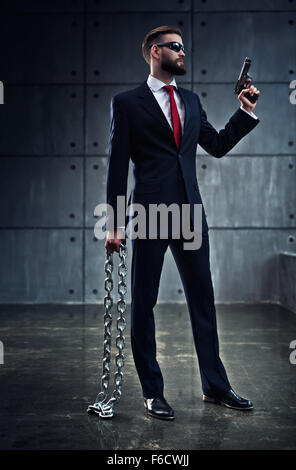 Danger businessman with heavy chain and gun in black suit standing on wall background. Stock Photo