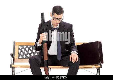 Depressed businessman holding a shotgun rifle and looking down seated on a wooden bench isolated on white background Stock Photo