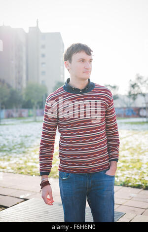 Knee figure of young handsome man overlooking left in the city, wearing blue jeans and a red striped sweater, hands in pocket - Stock Photo