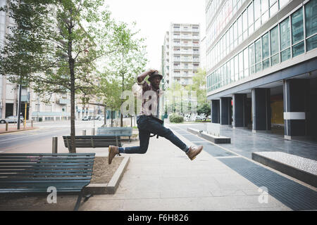 Young handsome afro black man jumping in the street of the city, looking upward surprised, holding his hat on his head - surprised, jumping, having fun concept Stock Photo