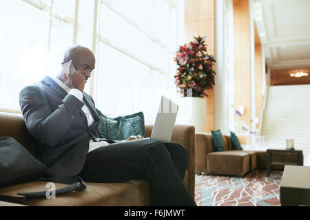 Businessman sitting on sofa working using cell phone and laptop. African male executive waiting in hotel lobby. Stock Photo
