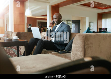 Happy young businessman sitting on sofa working using cell phone and laptop. African male executive waiting in hotel lobby. Stock Photo