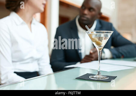 Close up shot of glass of cocktail on counter with two business people sitting in background discussing at cafe. Stock Photo