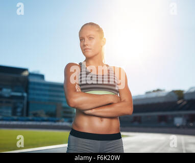 Portrait of fit young woman in sportswear standing on racetrack with her hands folded and looking away. Professional athlete in Stock Photo