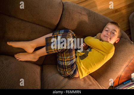 Looking down on a seven year old boy in pajamas lying on a sofa hugging his teddy bear, in Issaquah, Washington, USA Stock Photo