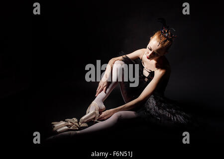 Slim ballerina in a black corset and black tutu tying pointe shoes. Classical Ballet. Photography in low key. Stock Photo