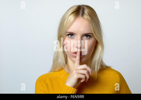 Close up Pretty Blond Woman Showing Shushing Gesture While Staring at the Camera Against Gray Background. Stock Photo