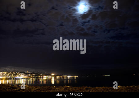 full moon over silent reflective fjord water with city lights in the background