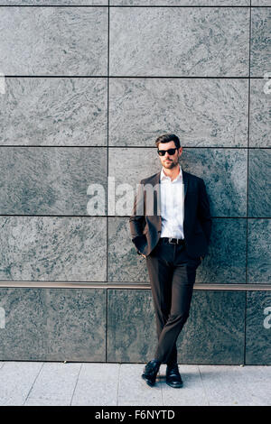 Young handsome modern businessman posing leaning against a wall, looking in camera, wearing a coat - business, success concept Stock Photo