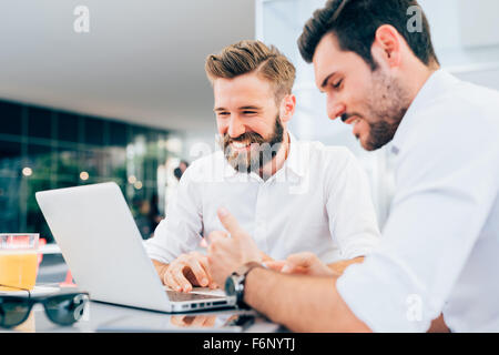 Two young bearded caucasian modern business man sitting in a bar, using laptop, looking downward, tapping on keyboard  - business, work, technology concept - focus on blonde hair man Stock Photo