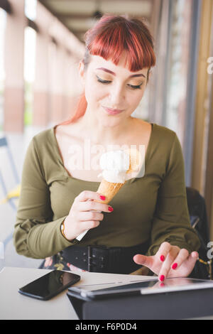 Young handsome caucasian redhead woman sitting in a bar using tablet while eating ice cream, looking downward - technology, social network, multitasking concept - wearing green shirt and floral skirt Stock Photo