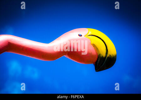 A detail of a cheeky, stylized and whimsical hand carved flamingo painted bright pink with yellow beak against a blue sky Stock Photo