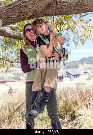 Attractive mother helps young son on rope swing hung from ranch tree Stock Photo