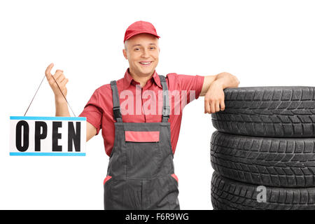 Young mechanic holding an open sign and leaning on a stack of car tires isolated on white background Stock Photo