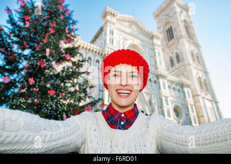 Smiling woman tourist in red knitted hat is taking selfie in front of traditional christmas tree near Duomo in Florence, Italy. Stock Photo