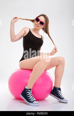 Young funny amusing cheerful sportswoman in black leotard and sneakers having fun with her hair sitting on pink fitball Stock Photo