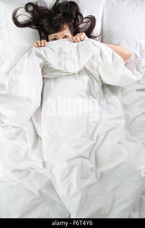 Mixed race woman peeking out from bed blankets Stock Photo