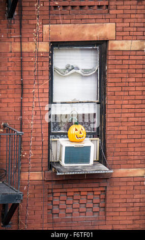 bright orange ceramic version hand carved Halloween pumpkin sitting on air conditioner outside window brick tenement building Stock Photo
