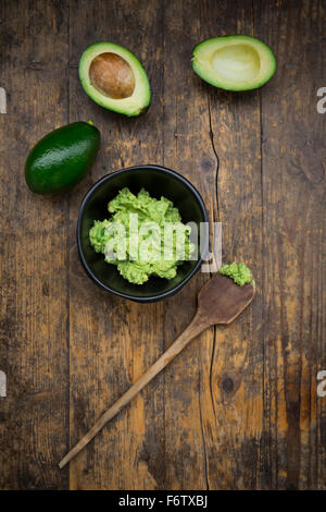 Bowl of Guacamole, cooking spoon and whole and sliced avocados on dark wood Stock Photo