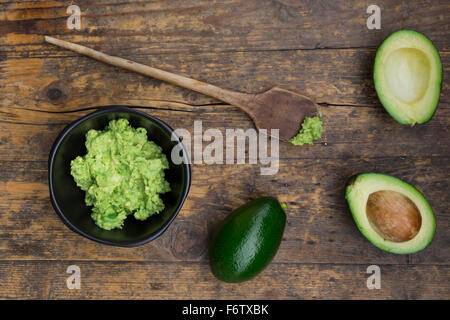 Bowl of Guacamole, cooking spoon and whole and sliced avocados on dark wood Stock Photo
