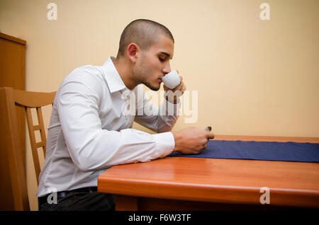Young Man Drinking from Espresso Coffee Cup, While Sitting at Table in his Home Kitchen Stock Photo