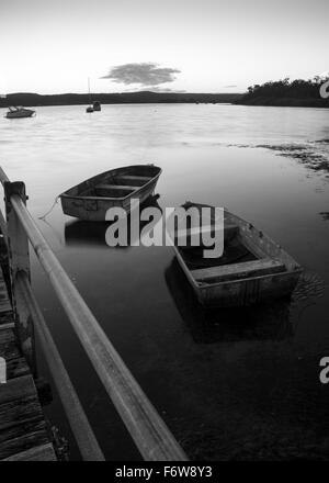 Two small open dinghy boats (tinnies) face the setting sun in calm water beside a wharf. monochrome Stock Photo