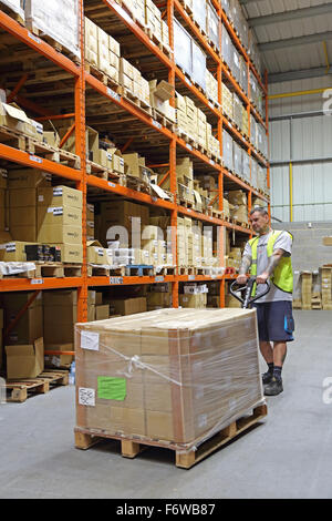 A worker pulls a pallet containing shrink-wrapped boxes in a modern storage warehouse with goods stacked on high racking Stock Photo