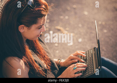 Young beautiful reddish brown caucasian woman seated on a sidewalk using a notebook - technology, communication, social network Stock Photo