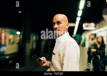 Young handsome caucasian bald business man standing on the underground platform, overlooking left waiting the tube holding a smartphone- work, waiting concept Stock Photo