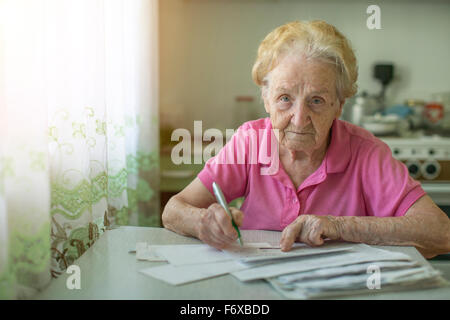 An elderly woman fills out a receipt for payment of utilities, sitting in the kitchen in house. Stock Photo