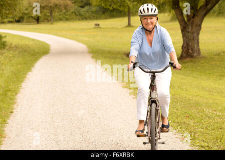 Fit healthy senior lady out cycling on her bicycle on a country road wearing a protective helmet and smiling happily at the came Stock Photo