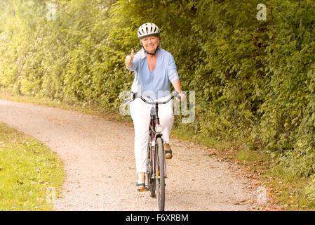 Fit healthy senior lady out cycling on her bicycle on a country road wearing a protective helmet and smiling happily at the came Stock Photo