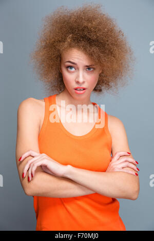 Beautiful curly wondered amazed surprised young woman in orange top shocked and posing with crossed arms on gray background Stock Photo