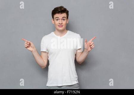 Confused indecisive doubtful handsome young man showing fingers in different directions over gray background Stock Photo