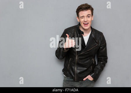 Portrait of a young man in leather jacket showing thumb up and winking on gray background Stock Photo