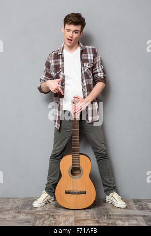 Full length portrait of a young man winking and pointing finger on guitar on gray background Stock Photo