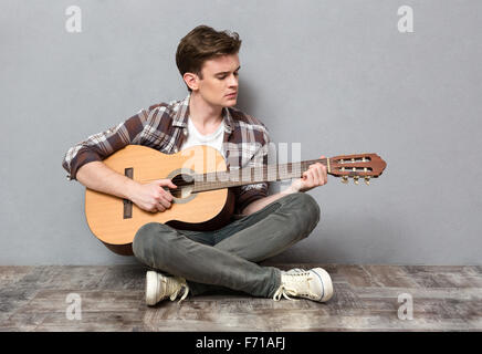 Portrait of a young man sitting on the floor and playing on guitar on gray background Stock Photo
