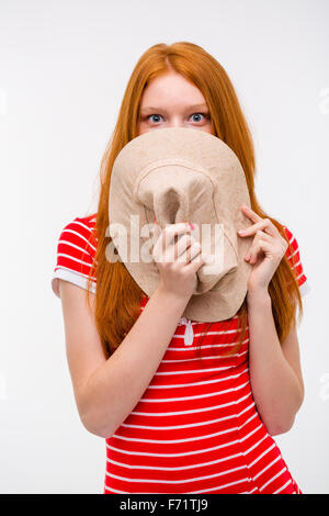 Embarrassed shy redhead young woman hiding her face behind hat posing on white background Stock Photo