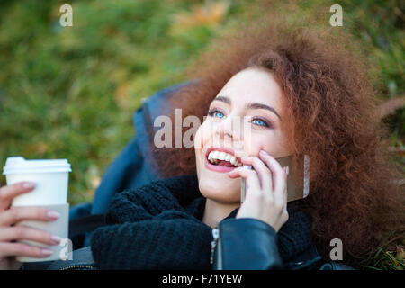 Portrait of a cheerful woman talking on the phone and holding cup with coffee while lying on green grass Stock Photo