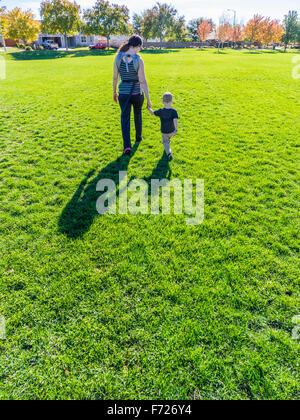 A mother holds the hand of her toddler son as they walk away together facing the other way and casting very long shadows. Stock Photo