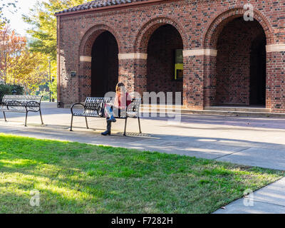 A female university student sits on a metal bench and studies in late afternoon light by Kendall Hall, CSU Chico, Chico, CA. Stock Photo