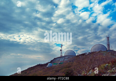 Three satellite dishes over on top of a mountain covered with protective domes on a background of clouds Stock Photo