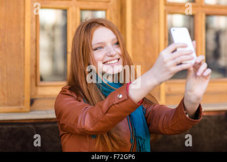 Happy smiling beautiful young woman with long red hair in leather jacket and scarf making selfie in outdoor cafe Stock Photo