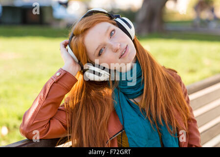 Portrait of relaxed charming young lady with beautiful long red hair enjoying music in headphones on bench in park Stock Photo